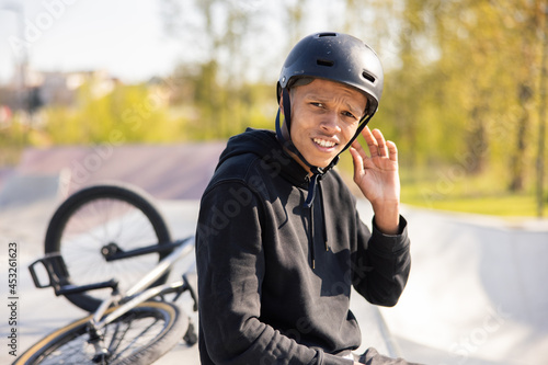 A young boy sits on a ramp at a skatepark with his bike lying wheel up behind him. The teenager cannot hear what his friends are saying to him, so he gestures with his hand near his ear