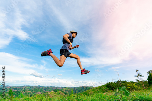 Asian man trail runners, wearing sportswear, are practicing running and jumping on the high mountain behind a beautiful view. There is a field of wind turbines generating electricity in the background