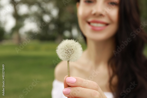 Beautiful young woman holding dandelion outdoors, focus on hand with flower. Allergy free concept