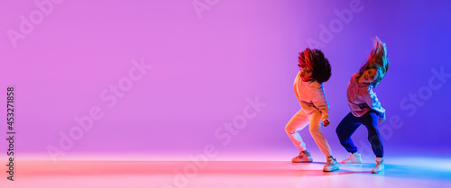 Two beautiful young girls dancing hip-hop dynamically on colorful gradient background in neon.