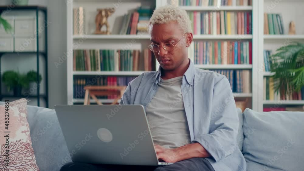 Happy young African American male sitting on couch in comfy pose using laptop screaming yes raising fist. Excited man celebrate victory in online game receive money reward for good job on bank account Happy young African American male sitting on couch in comfy pose using laptop screaming yes raising fist. Excited man celebrate victory in online game receive money reward for good job on bank account