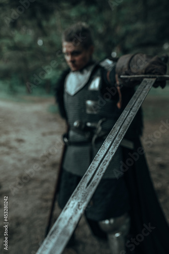 Medieval knight in armour stands among forest with sword in his hand.