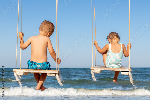 Back view couple of cute little adorable small caucasian sibling kids enjoy having fun swinging and playing at playground sea sand beach against blue ocean water wave and clear sky. Family vacation