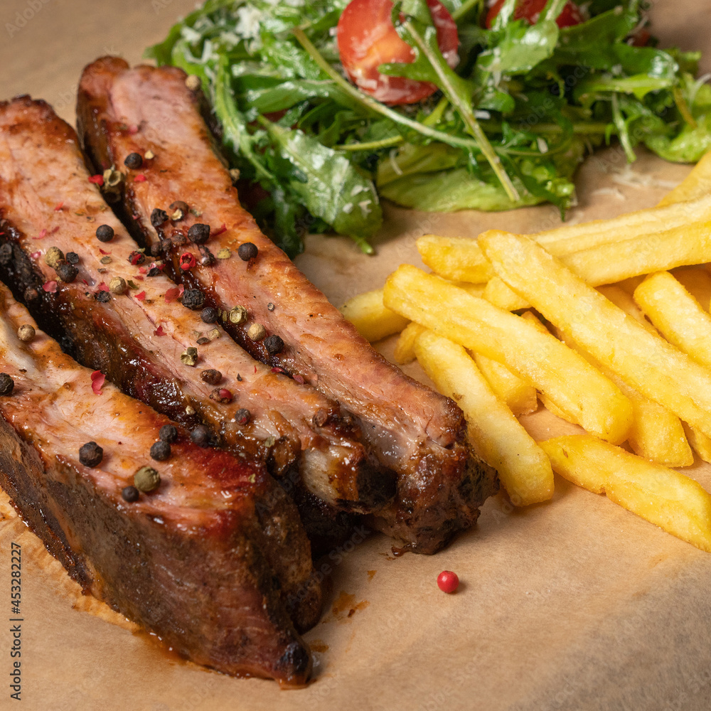 BBQ spare ribs served with French fries and arugula salad with cherry tomatoes and parmesan cheese served on board covered with food grade paper. Restaurant concept. Square cropped image. BBQ spare ribs served with French fries and arugula salad with cherry tomatoes and parmesan cheese served on board covered with food grade paper. Restaurant concept. Square cropped image.
