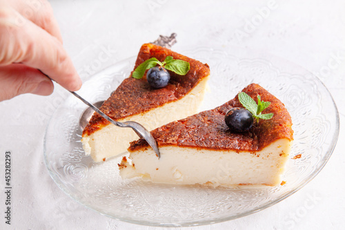A woman's hand takes a piece of cheesecake with a spoon. Slice of Homemade Basque burnt cheesecake with blueberries on light background.