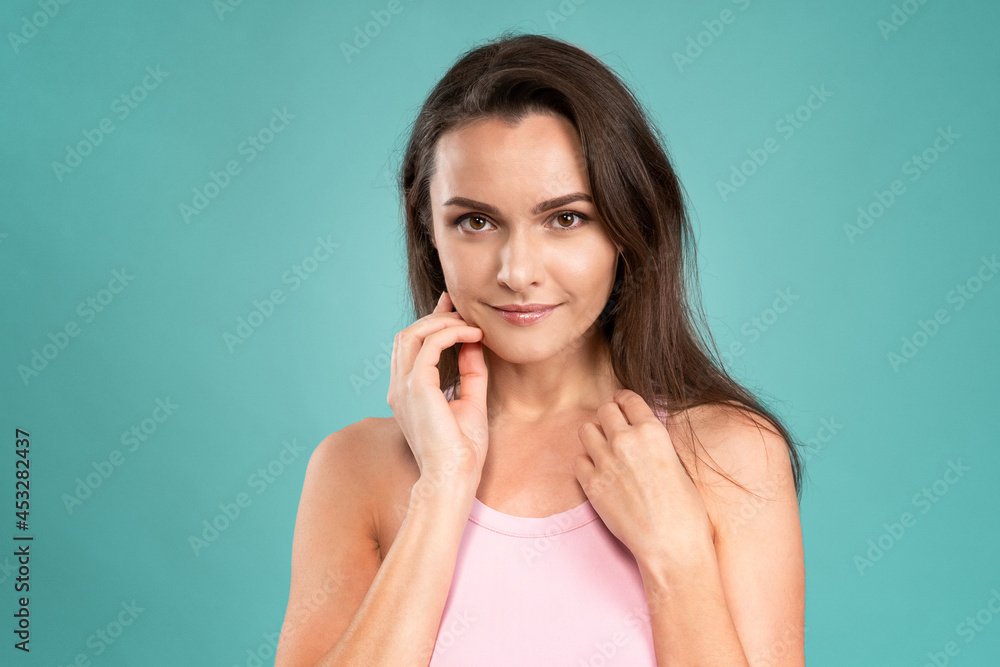 Fototapeta premium No makeup beautiful woman moisturising her face standing with hands on her neck and chicks, massaging it gently attractive brunette girl on blue background. Skin care concept. 