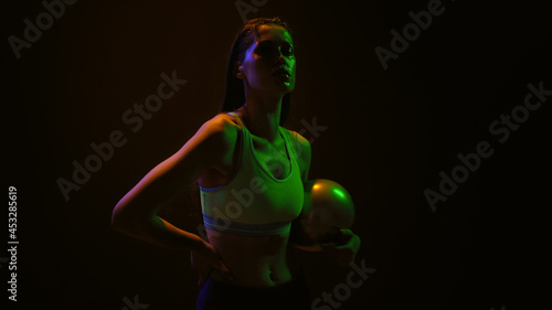 Beautiful girl posing holding a ball in hands standing in neon lights wearing sport top stretching neck muscles turning head on black background. 
