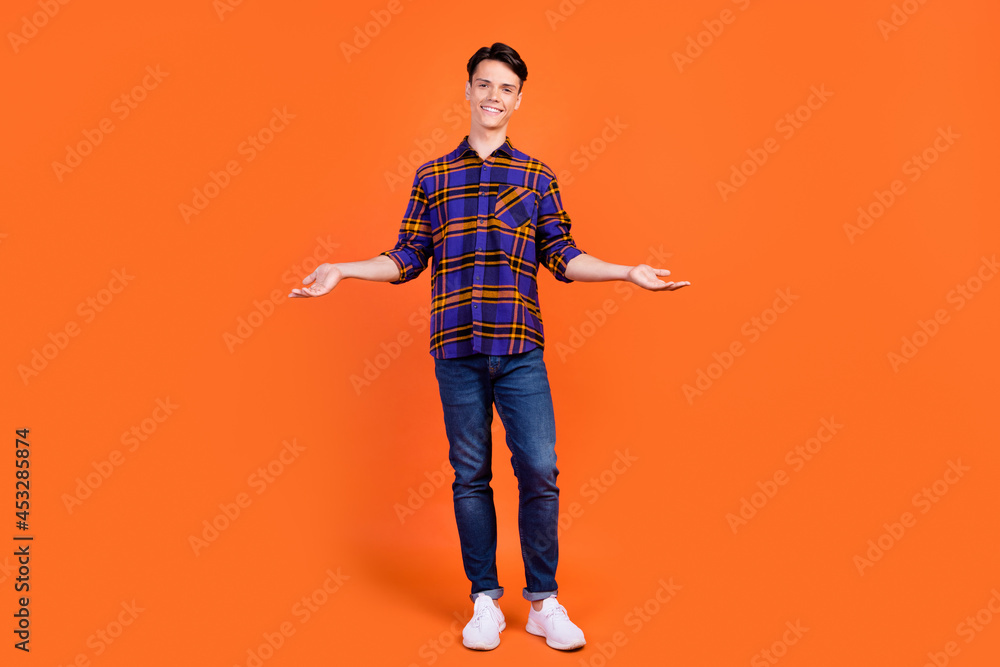 Photo of sweet cute teen boy wear plaid outfit spectacles smiling compare arms empty space isolated orange color background Photo of sweet cute teen boy wear plaid outfit spectacles smiling compare arms empty space isolated orange color background