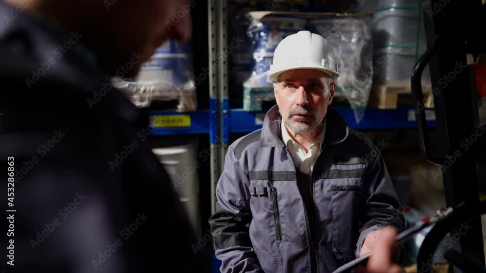 Professional bearded senior freight agent arguing with warehouse loader in storage indoors pointing at documents. Dissatisfied confident professional man scolding worker for failure indoors. Conflict Professional bearded senior freight agent arguing with warehouse loader in storage indoors pointing at documents. Dissatisfied confident professional man scolding worker for failure indoors. Conflict