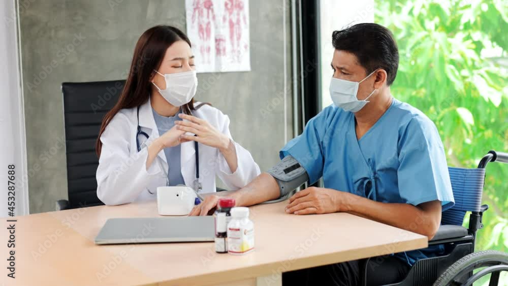 Asian woman doctor with face mask checks stethoscope health of an elderly handicapped man in a wheelchair by a blood pressure monitor at a hospital clinic.Older people healthcare concept. Asian woman doctor with face mask checks stethoscope health of an elderly handicapped man in a wheelchair by a blood pressure monitor at a hospital clinic.Older people healthcare concept.