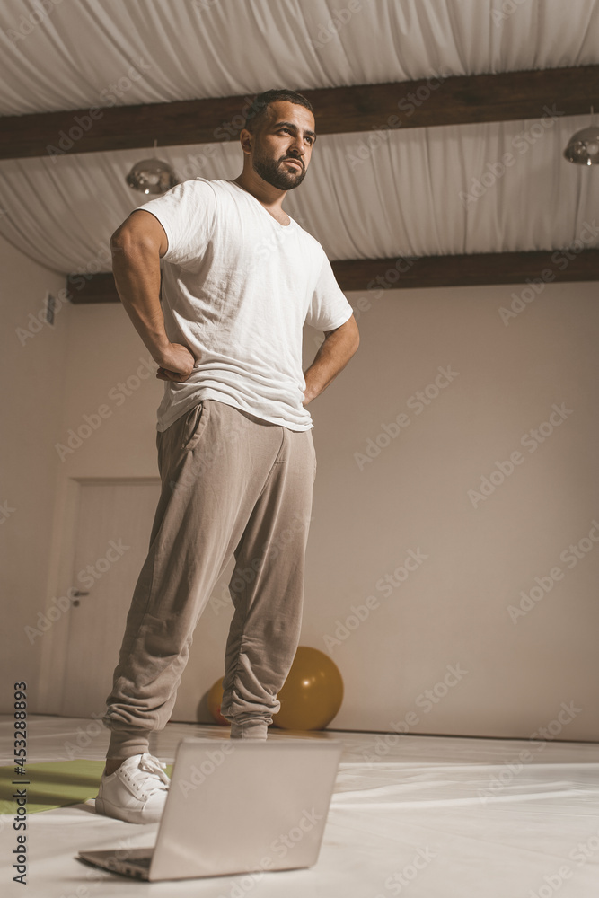 sportive young man trains hard at home standing in front of laptop. Self isolated and motivated young man warm-up doing special exercises for muscles. Work out during coronavirus lockdown. sportive young man trains hard at home standing in front of laptop. Self isolated and motivated young man warm-up doing special exercises for muscles. Work out during coronavirus lockdown.