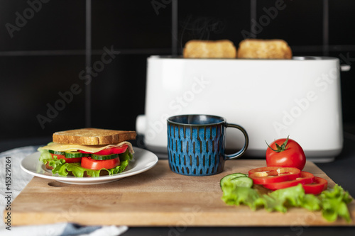 Delicious and healthy breakfast. Tasty sandwich on white plate, blue cup of hot tea or coffee with steam, fresh tomatoes and salad on wooden board. On background white toaster with bread. Soft focus.