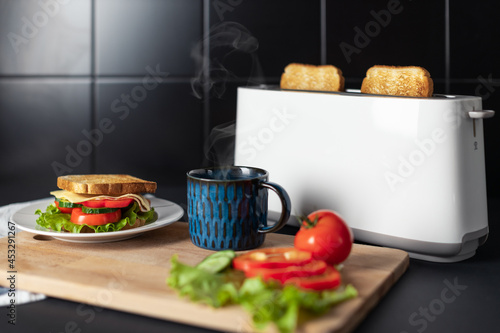 Toasted bread in white toaster. On wooden board blue cup of hot tea with steam, fresh tomatoes, salad and ready made sandwich. Black tile on background. Ingredients for delicious and healthy breakfast