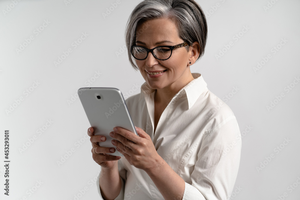 Portrait of a grey haired senior woman in white shirt and eye glasses using digital tablet. Technology by the elderly browsing internet shopping online using pad isolated on white background. Portrait of a grey haired senior woman in white shirt and eye glasses using digital tablet. Technology by the elderly browsing internet shopping online using pad isolated on white background.