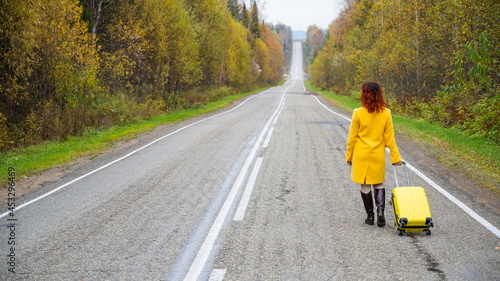 A red-haired woman in a yellow coat walks with a yellow suitcase along the highway in autumn.
