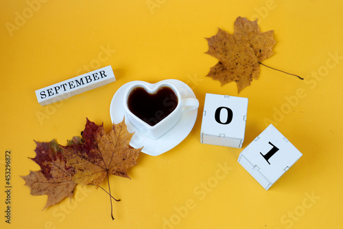 Calendar for September 1 : the name of the month in English, cubes with the numbers 0 and 1,a white heart-shaped cup on a saucer with tea, maple leaves, yellow background, top view photo