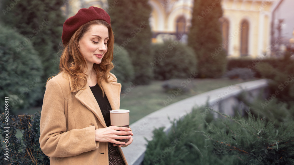 Beautiful young woman holding a mug with coffee standing outdoors. Portrait of stylish young woman wearing autumn coat and red beret outdoors. Autumn accessories. Beautiful young woman holding a mug with coffee standing outdoors. Portrait of stylish young woman wearing autumn coat and red beret outdoors. Autumn accessories.