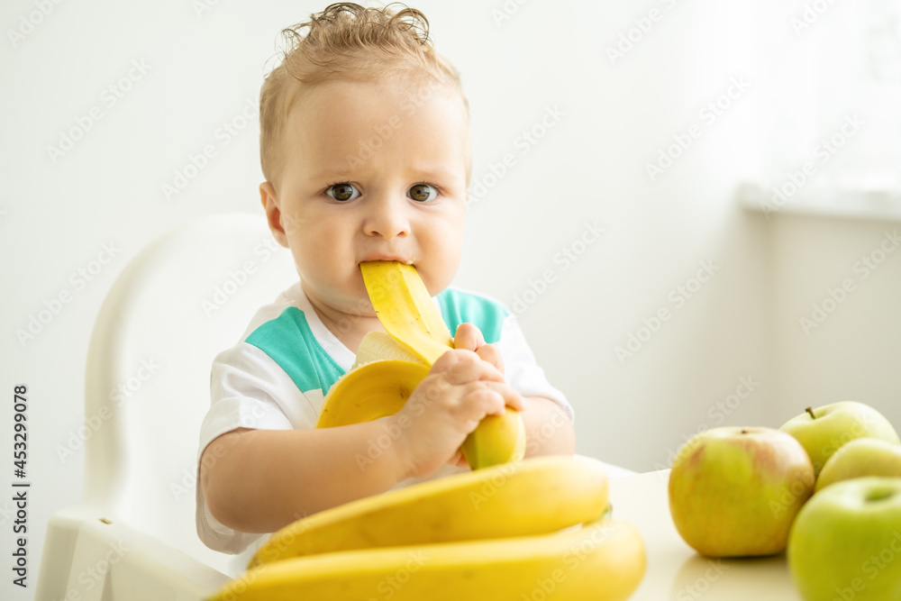 funny baby boy sitting at the table in child chair eating banana on white kitchen. funny baby boy sitting at the table in child chair eating banana on white kitchen.