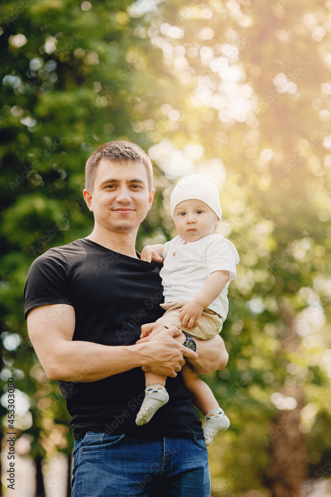 Fototapeta premium Portrait happy child boy sitting on hand of young dad, against background of green grass in park