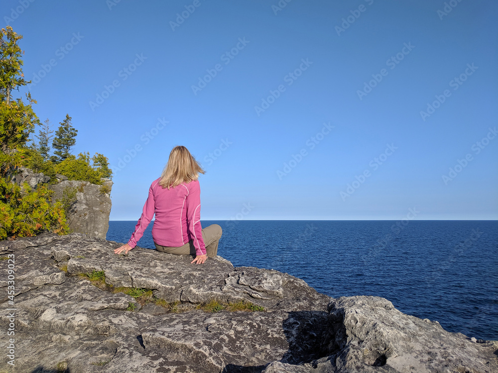 Young woman sits on a high rocky cliff edge overlooking the sea on a sunny day. Bruce Peninsula National Park, Georgian Bay, Ontario, Canada. Hiking, camping, active lifestyle concept. Young woman sits on a high rocky cliff edge overlooking the sea on a sunny day. Bruce Peninsula National Park, Georgian Bay, Ontario, Canada. Hiking, camping, active lifestyle concept.