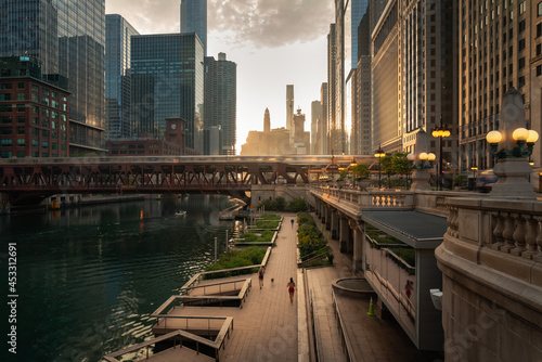 Beautiful downtown Chicago morning along the river as people jog on the path below and train crosses a bridge as the sun casts yellow light into the scene from behind the high-rise buildings beyond.