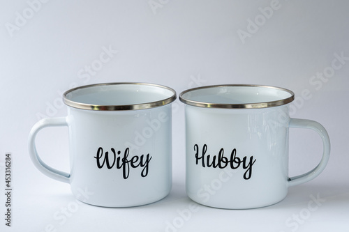 Two white enamel mugs with writings "Hubby" and "Wifey"on them on a white background. Wedding accessories, cups for coffee or tea, luxury wedding.