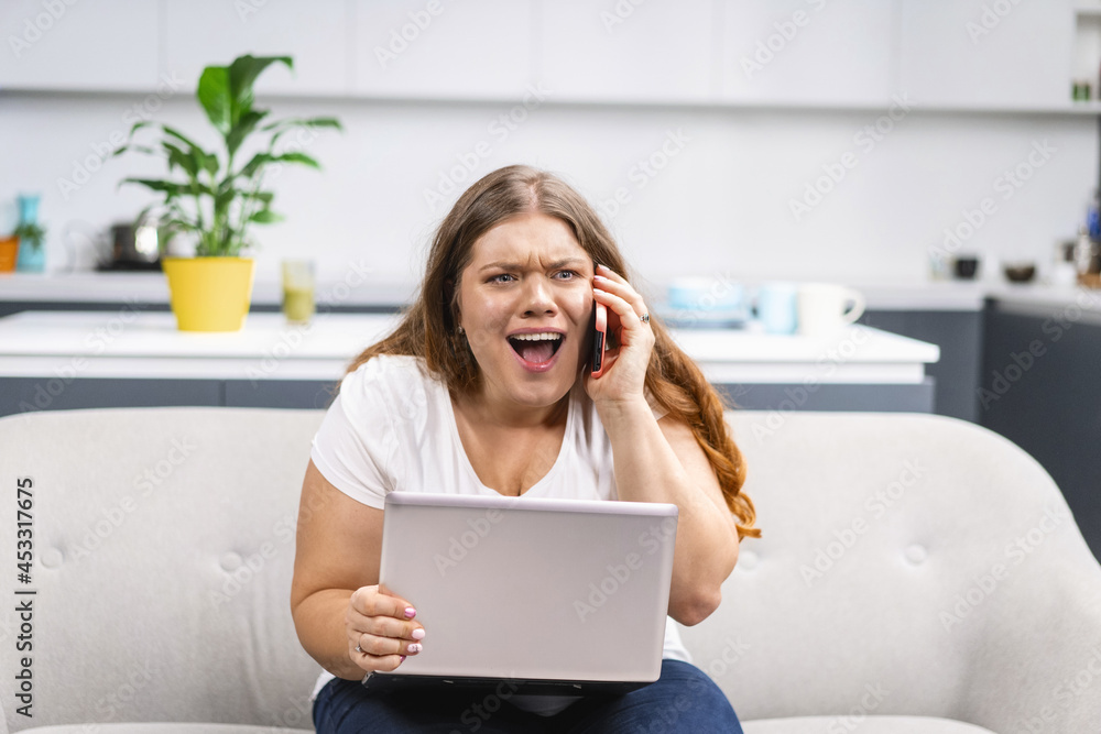 Mad screaming talking on the phone young fat girl working on laptop sitting on the sofa at home with the kitchen on background. Self acceptance. Working distantly from home using laptop. Mad screaming talking on the phone young fat girl working on laptop sitting on the sofa at home with the kitchen on background. Self acceptance. Working distantly from home using laptop.
