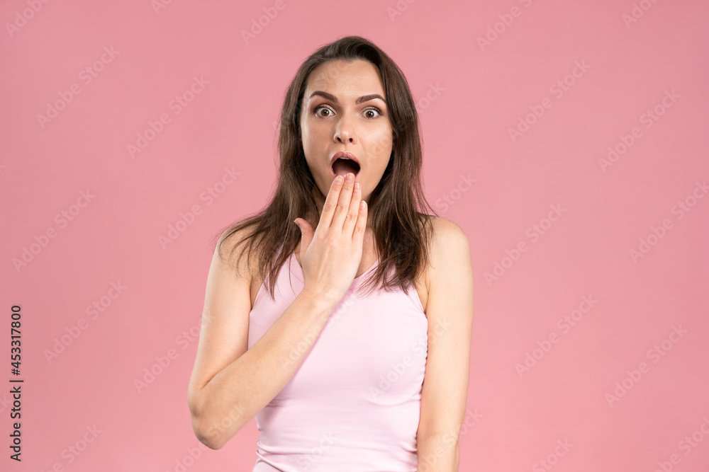 Astonished young woman looking surprised with dark hair in pink t-shirt looking at camera with holding hand at the lips. Facial expressions, emotions, feelings. Astonished young woman looking surprised with dark hair in pink t-shirt looking at camera with holding hand at the lips. Facial expressions, emotions, feelings.