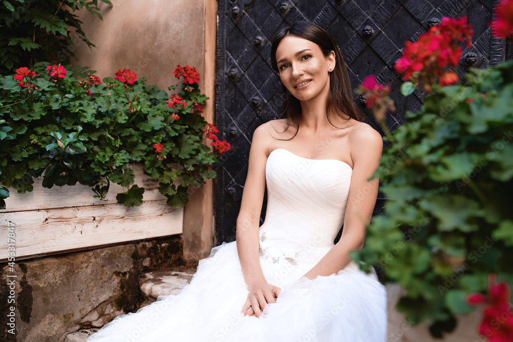 Beautiful caucasian brunette bride sitting among flowers. Wedding day in old beautiful European city.