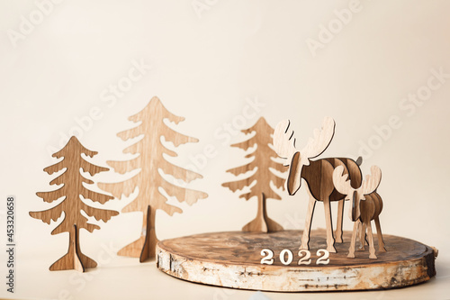 Christmas handmade reindeer, fir trees and numbers 2022 on wooden stand. New year holiday greeting card. Christmas decorative figure deer on pastel beige background. Wooden vintage and eco style. © Lyubov