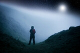 A concept edit of a lone hooded hiker  on a path in the countryside looking up on a cold spooky, foggy night with stars above