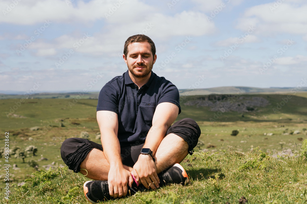 Relaxed bearded man with closed eyes sitting in meadow and meditating while establishing connection with nature in a sunny morning