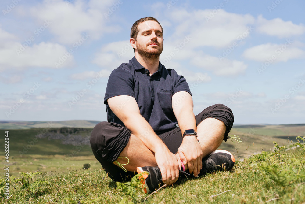 Relaxed bearded man with closed eyes sitting in meadow and meditating while establishing connection with nature in a sunny morning