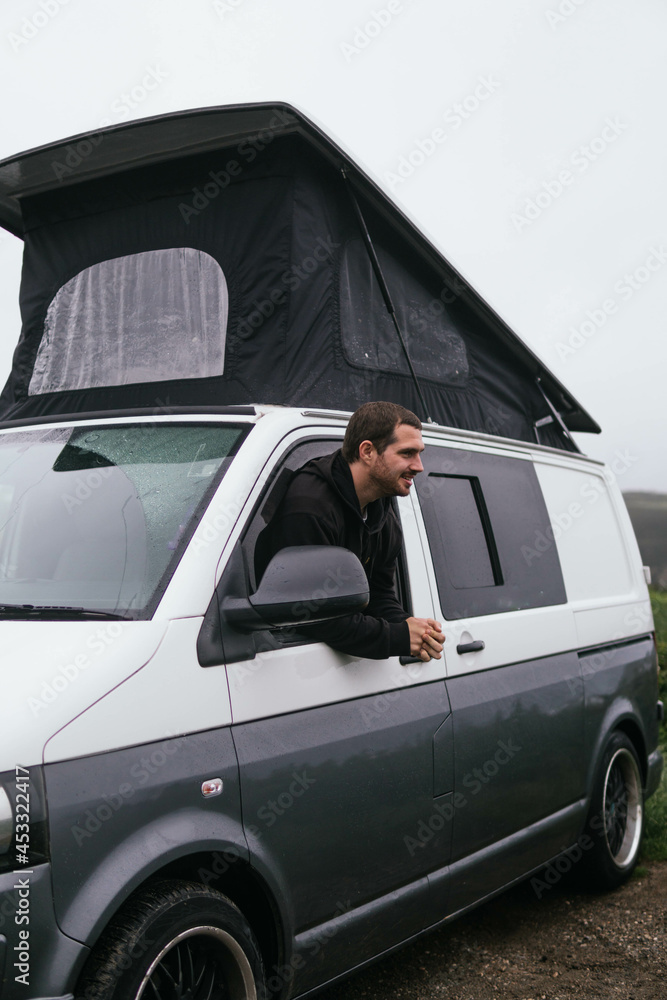 Smiling young guy peeking out of opened window of camper van and looking at camera while traveling during winter vacation in UK