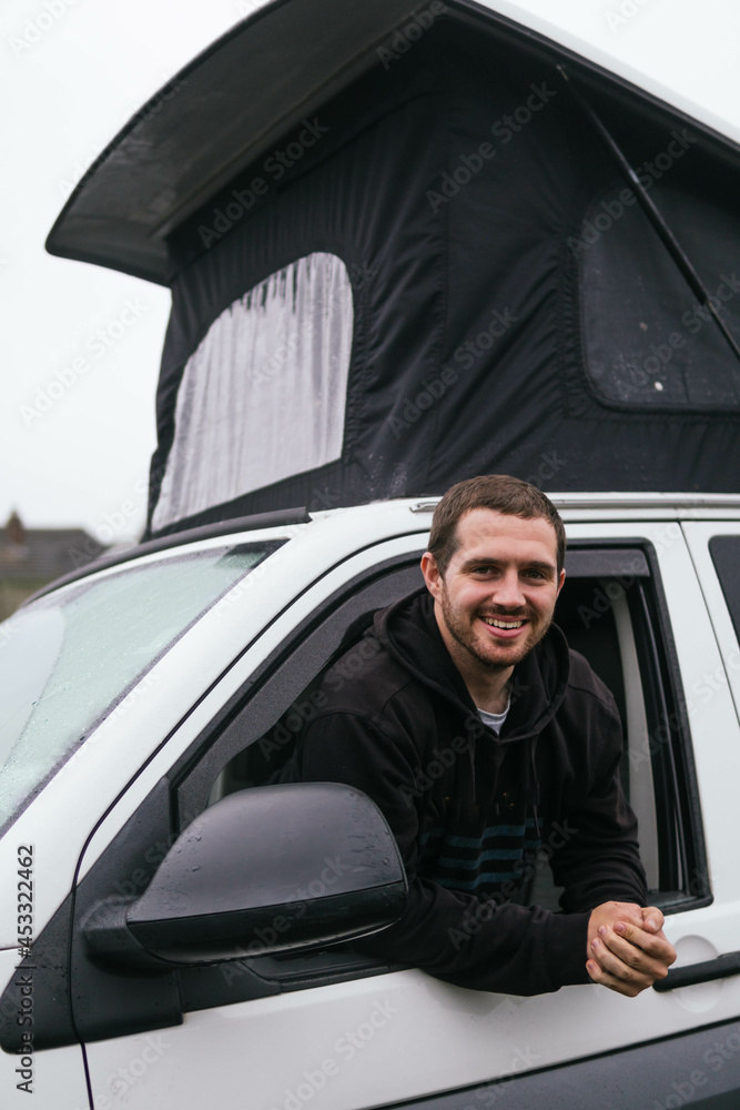 Smiling young guy peeking out of opened window of camper van and looking at camera while traveling during winter vacation in UK