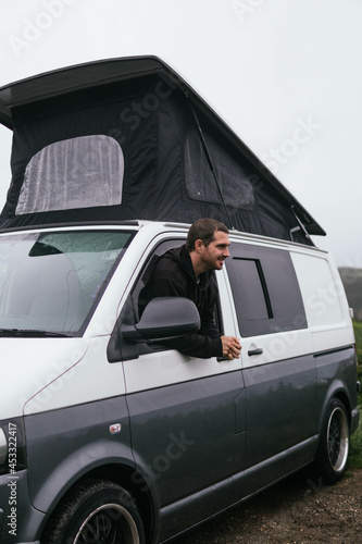 Obraz na płótnie Smiling young guy peeking out of opened window of camper van and looking at camera while traveling during winter vacation in UK w salonie
