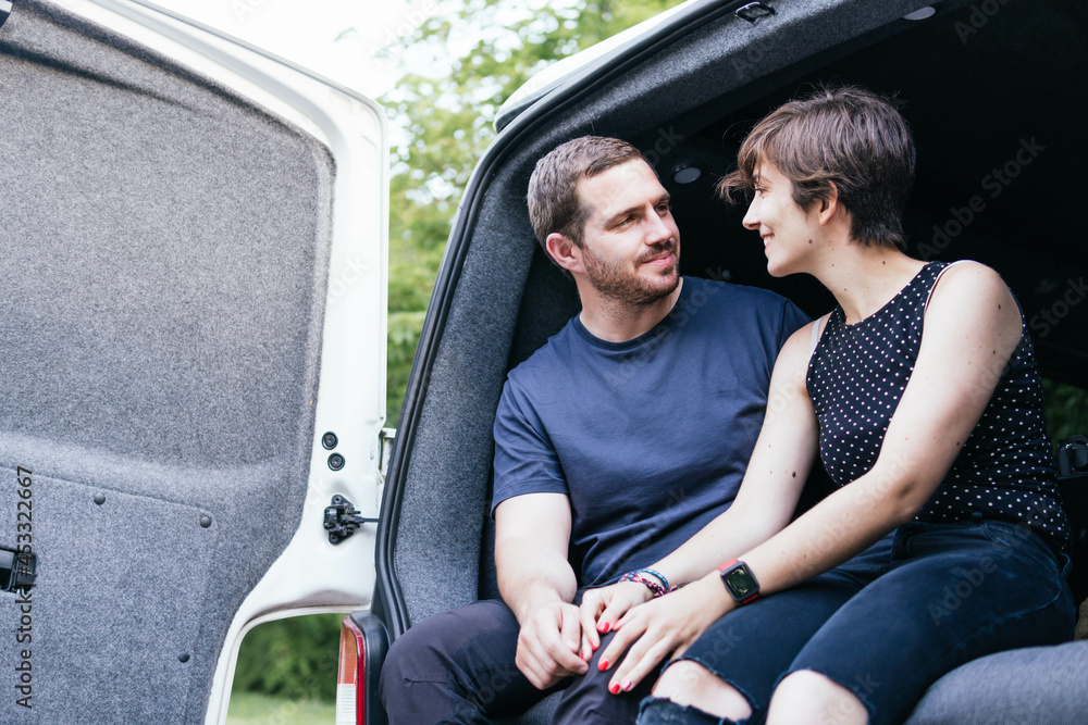 Young couple sitting on a camper van looking at each other and enjoying a good time while travelling. Enjoying simple pleasures.