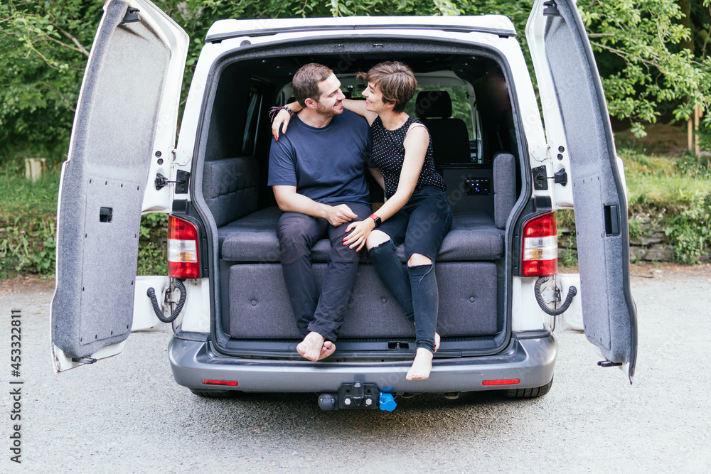 Young couple sitting on a camper van looking at each other and enjoying a good time while travelling. Enjoying simple pleasures.