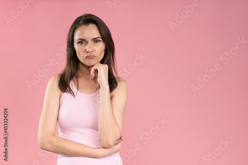 Annoyed or bothered young woman wearing pink t-shirt holding hands folded with chin leaned on one, Female half-length portrait. Human emotions, facial expression concept.