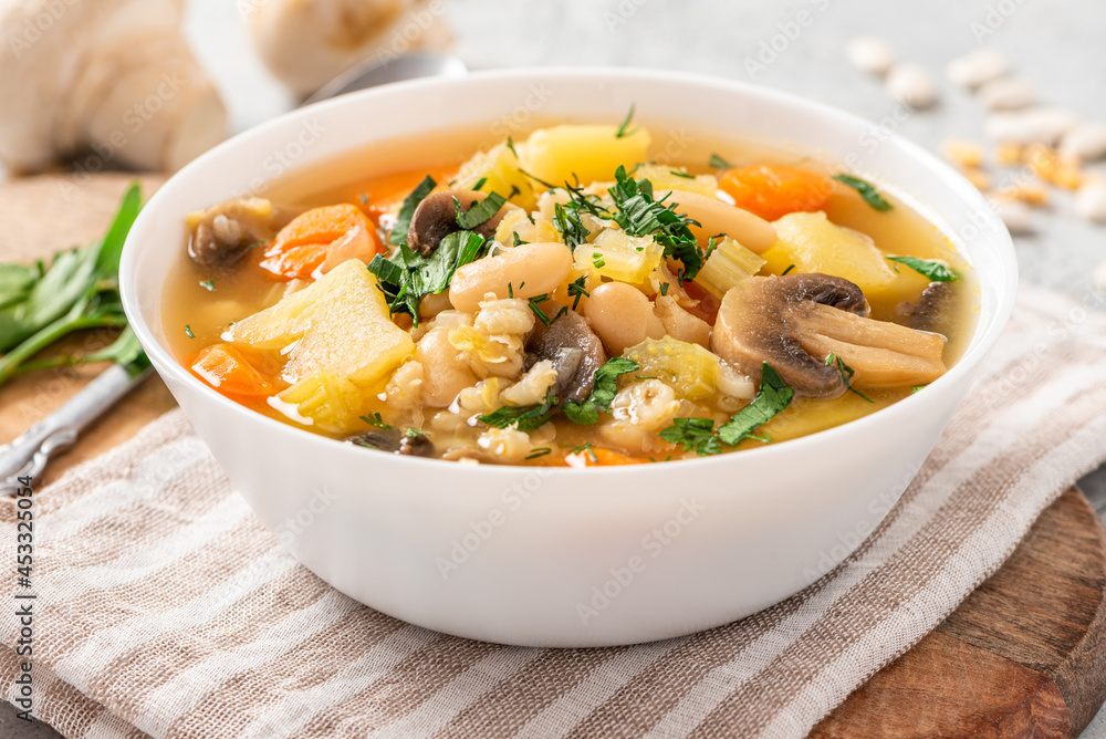 Soup with barley, beans, mushrooms, potatoes, carrots, celery, and herbs. Barley soup with vegetables in a white bowl on a gray concrete background. Vegan food, homemade soup. Soup with barley, beans, mushrooms, potatoes, carrots, celery, and herbs. Barley soup with vegetables in a white bowl on a gray concrete background. Vegan food, homemade soup.