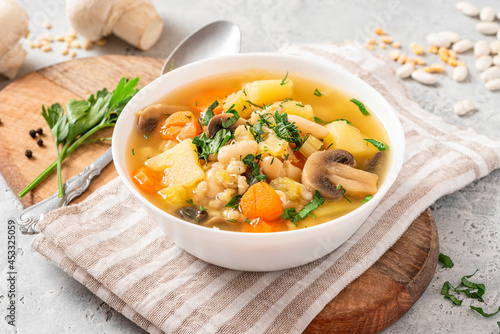 Soup with barley, beans, mushrooms, potatoes, carrots, celery, and herbs. Barley soup with vegetables in a white bowl on a gray concrete background. Vegan food, homemade soup.