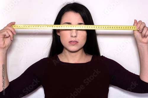 Mujer joven morena con cabello largo sosteniendo una cinta métrica amarilla a la altura de sus ojos cubriendo su vista sobre un fondo blanco.