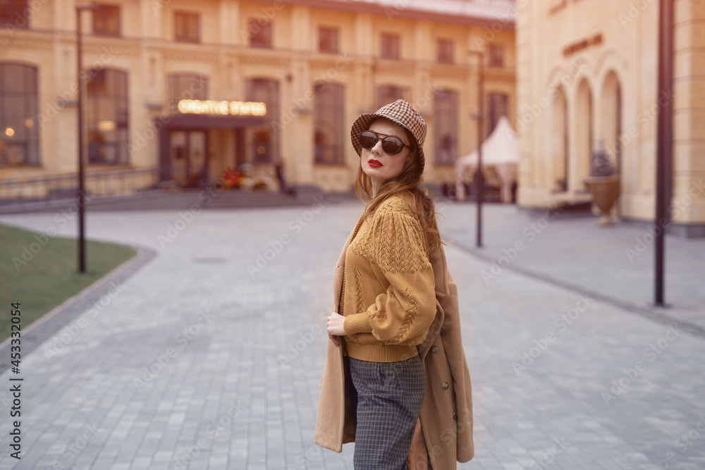 Turned from behind fashion model young woman in sunglasses and hanging on a shoulder autumn beige coat walking on the street happily posing for the camera. Tinted photo. Turned from behind fashion model young woman in sunglasses and hanging on a shoulder autumn beige coat walking on the street happily posing for the camera. Tinted photo.