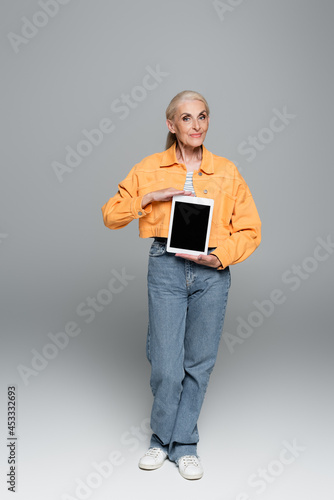 elderly woman in trendy clothes showing digital tablet with blank screen on grey