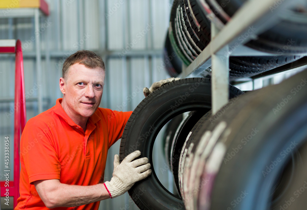 Car service. Male worker checking new tire wheel on shelves shelf at wheel store, wearing red uniform, gloves during working. Specialist technician worker examining new tire wheel. Maintenance concept Car service. Male worker checking new tire wheel on shelves shelf at wheel store, wearing red uniform, gloves during working. Specialist technician worker examining new tire wheel. Maintenance concept