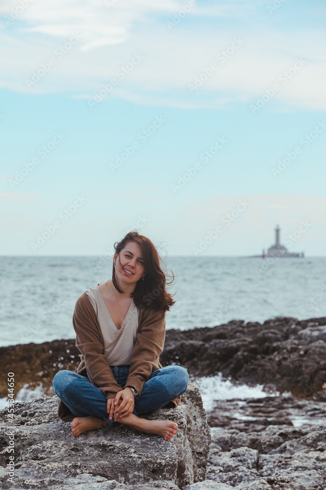 woman sitting by rocky sea beach in wet jeans lighthouse on background