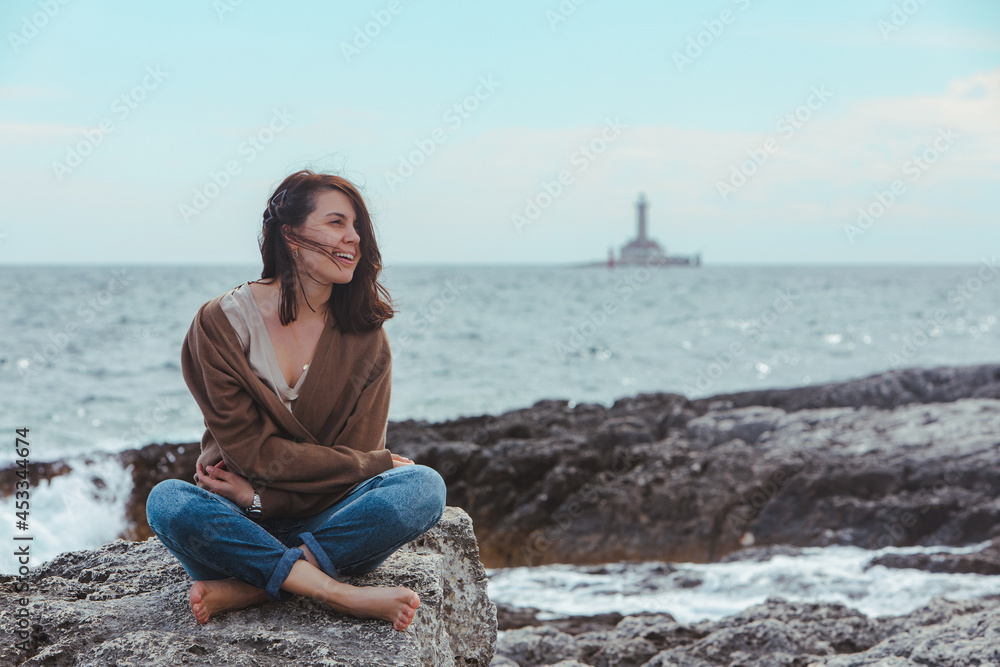 woman sitting by rocky sea beach in wet jeans lighthouse on background