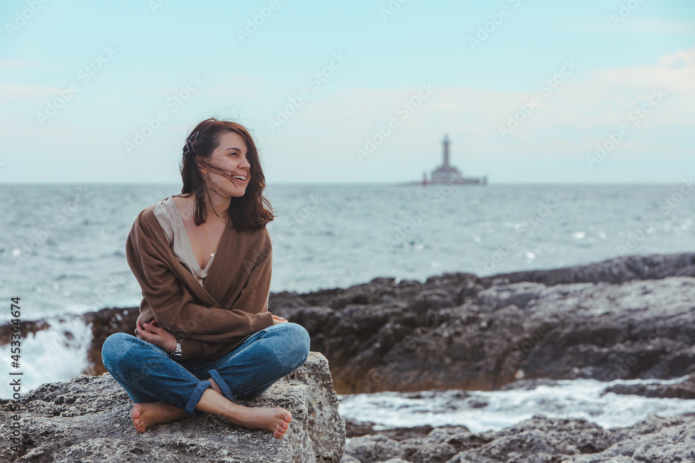 woman sitting by rocky sea beach in wet jeans lighthouse on background woman sitting by rocky sea beach in wet jeans lighthouse on background