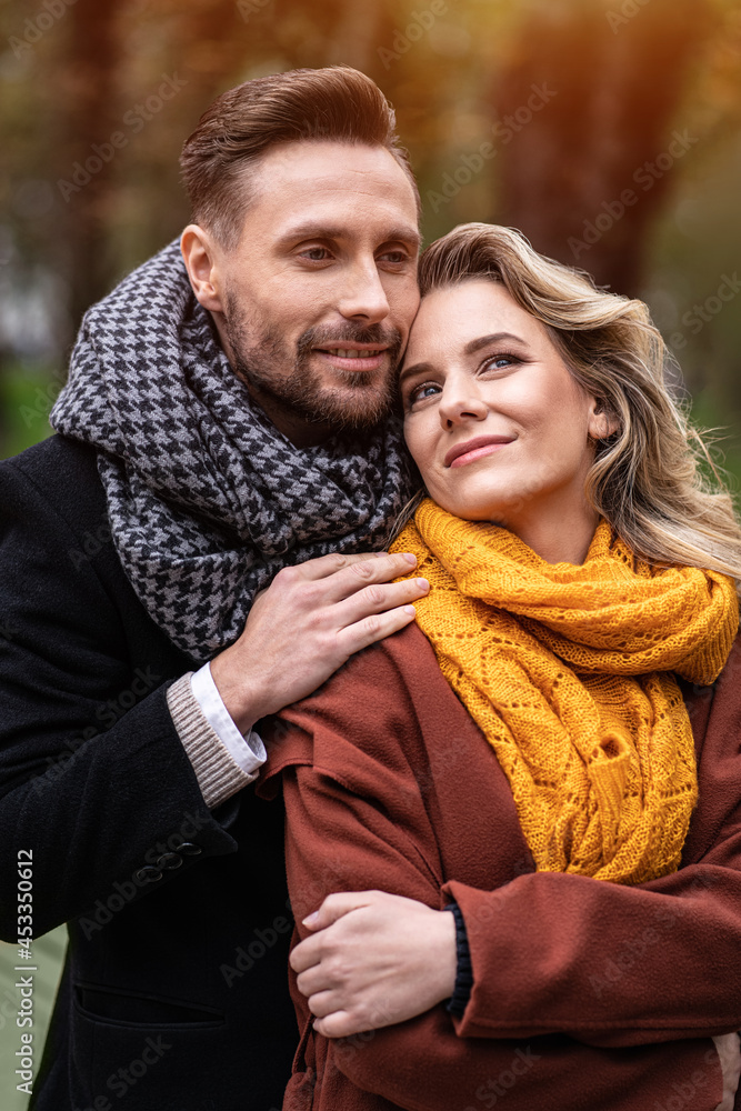 Fototapeta premium Tenderness of a cute couple. Handsome man and a woman hugged from behind smile looking at each other in the autumn park. Outdoor shot of a young couple in love having great time.