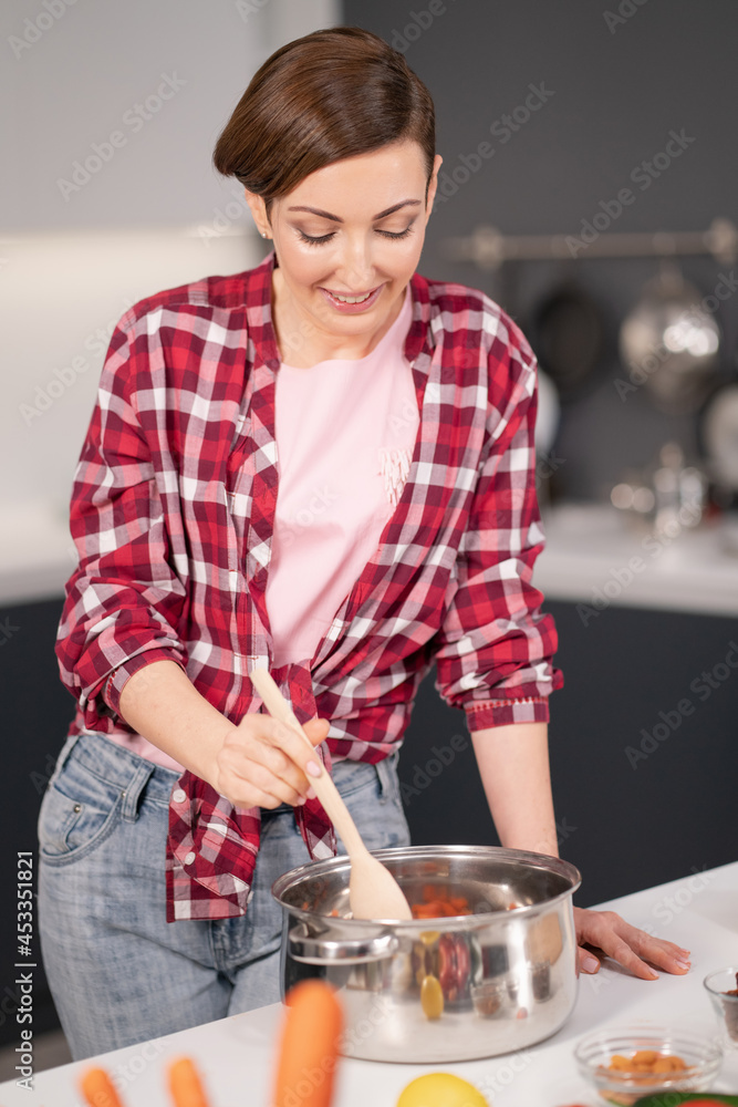 Woman mixing and trying a cooked dish in a pan using a long wooden spoon. Woman with a short hair cooking a dinner for family standing in the modern kitchen of a new house. Healthy food at home. Woman mixing and trying a cooked dish in a pan using a long wooden spoon. Woman with a short hair cooking a dinner for family standing in the modern kitchen of a new house. Healthy food at home.